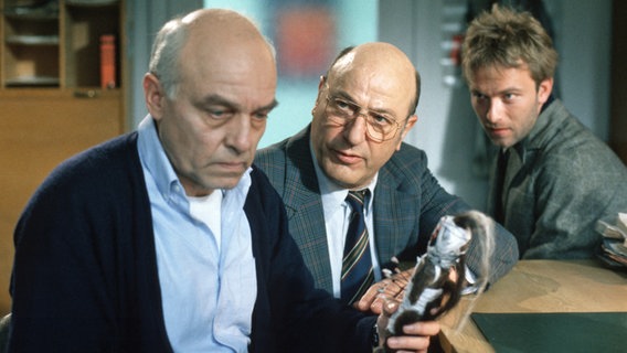 Kommissar Stoever (Manfred Krug, Mitte) und Brockmöller (Charles Brauer, links) gehen gerade noch einmal die Beweislage im Mordfall des Unternehmers Walter Mewes durch, als Robbie (Oliver Betke, rechts) einen angsteinflößenden Fund abliefert. Eine Vodoopuppe mit Brockmöllers Foto. Der Kommissar starrt mit angsterfülltem Blick auf dem Figur, denn genauso eine wurde von dem Mordopfer Walter Mewes gefunden. Kommissar Stoever ist irritiert. © © NDR/Studio HH 