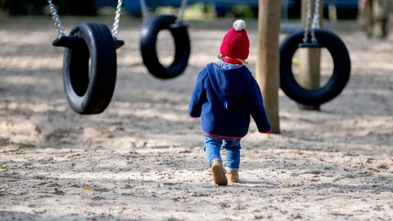 Blick von hinten auf ein kleines Kind, das auf einem Spielplatz zu Schaukelreifen läuft. © dpa Foto: Christoph Soeder