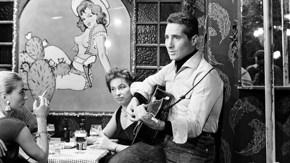 Szene aus der Stahlnetz-Folge "Die Tote im Hafenbecken" von 1958: Freddy Quinn spielt Gitarre. Zwei Damen sitzen daneben am Tisch. © NDR 
