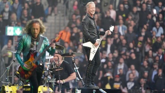 Sänger James Hetfield, Schlagzeuger Lars Ulrich und Gitarrist Kirk Hammett von Metallica stehen auf der Bühne im Hamburger Volksparkstadion. © dpa Foto: Marcus Brandt