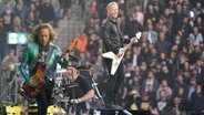 Sänger James Hetfield, Schlagzeuger Lars Ulrich und Gitarrist Kirk Hammett von Metallica stehen auf der Bühne im Hamburger Volksparkstadion. © dpa Foto: Marcus Brandt