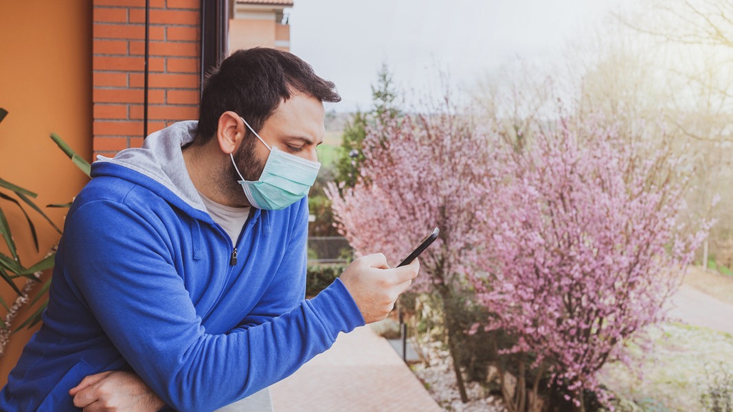 Ein Mann steht mit einer Maske vor dem Mund auf einem Balkon und schaut auf sein Handy. 