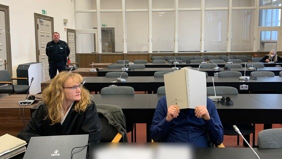 Ein Angeklagter, der mehrere Biomärkte in Hamburg überfallen haben soll, sitzt neben seiner Verteidigerin mit Mappe vorm Gesicht in einem Saal des Landgerichts. © Elke Spanner 