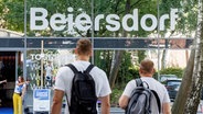 Der Schriftzug "Beiersdorf" ist über dem Eingang der neuen Konzernzentrale in Hamburg zu lesen. Davor sind zwei Menschen zu sehen, die Richtung Eingang gehen. © picture alliance / dpa Foto: Markus Scholz