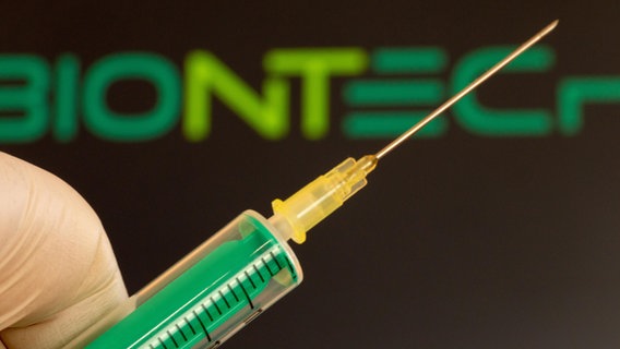 Nahaufnahme einer Injektionsnadel, im Hintergrund das Logo von Biontech. © picture alliance / CHROMORANGE | Udo Herrmann 