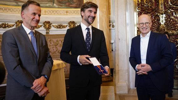 Hochseesegler Boris Herrmann steht mit seinem Bundesverdienstkreuz zwischen Sportstaatsrat Christoph Holstein und Bürgermeister Peter Tschentscher (beide SPD). © Christian Charisius/dpa 