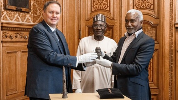 Hamburgs Kultursenator Carsden Brosda (l.), Abba Isa Tijani, Generaldirektor der Nationalen Behörde für Museen und Denkmäler in Nigeria, und Yusuf Matama Tuggur, Botschafter der Bundesrepublik Nigeria, bei der Übergabe einer Benin-Bronze an die Republik Nigeria. © picture alliance / dpa Foto: Markus Scholz