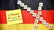 Buchstaben bilden die Schriftzüge "Einheit" und "Deutschland" auf einer deutschen Fahne. Darauf liegt auch ein mit Notizzettel mit der Aufschrift "3. Oktober - Tag der Deutschen Einheit". © picture alliance / CHROMORANGE Foto: Christian Ohde
