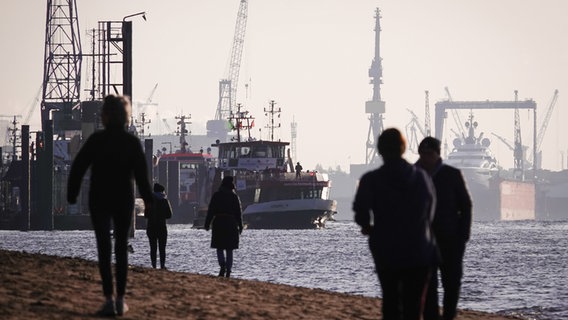 Menschen gehen am am Elbstrand von Övelgönne spazieren. Im Hintergrund ist der Hamburger Hafen zu sehen. © picture alliance / dpa Foto: Christian Charisius