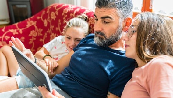 Ein Vater sitzt mit seinen Töchtern auf dem Sofa und schaut auf ein Tablet. © imago images/Giorgio Magini Foto: Giorgio Magini