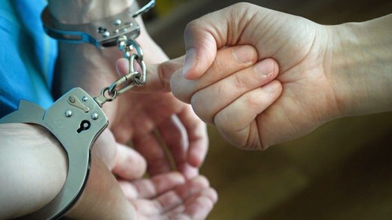Symbolbild: Mit dem Zeigefinger hakt sich eine Person in die Kette der Handschellen ihres Gefangenen, um ihn wegzuführen. © dpa/picture alliance Foto: Hans Wiedl/dpa-Zentralbild