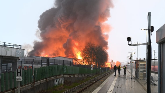 Polizisten stehen an einer Haltestelle am Bahnhof Hamburg-Rothenburgsort, während Flammen eines Großbrandes im Hintergrund zu sehen sind. © Jonas Walzberg/dpa 