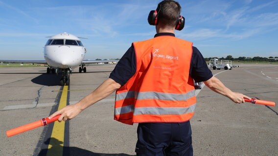 Ein Flughafen-Mitarbeiter weist auf dem Flughafen Hamburg eine Maschine auf dem Vorfeld ein. © picture-alliance Foto: Christian Charisius