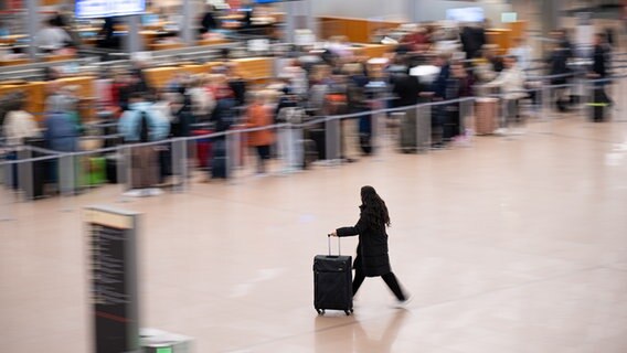 Eine Frau schiebt ihren Koffer durch das Terminal 1 am Hamburger Flughafen an einer Schlange wartender Reisender vorbei. © picture alliance/dpa | Daniel Reinhardt Foto: Daniel Reinhardt