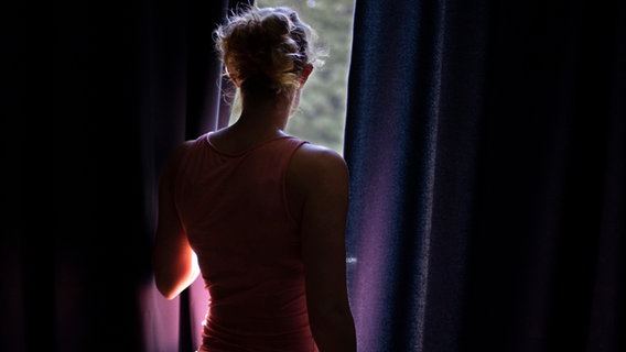 Eine junge Frau steht vor einem Fenster und guckt durch den Vorhang nach draußen. © photothek Foto: Ute Grabowsky