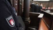 Die Rechtsanwälte stehen vor den angeklagten Frauen im Sitzungssaal im Strafjustizgebäude zu Beginn des Prozesses gegen eine Familie des mutmaßlichen Drahtziehers bei Schüssen auf ein Rockergang-Mitglied. Im Vordergrund ist ein Arm eines Justizbeamten zu sehen. © picture alliance / dpa Foto: Marcus Brandt