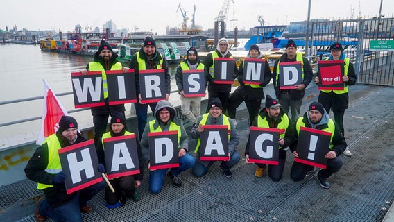 Streikende stehen mit Warnwesten und Fahnen vor stillliegenden Fähren am Hadag-Anleger. © picture alliance/dpa Foto: Jonas Walzberg