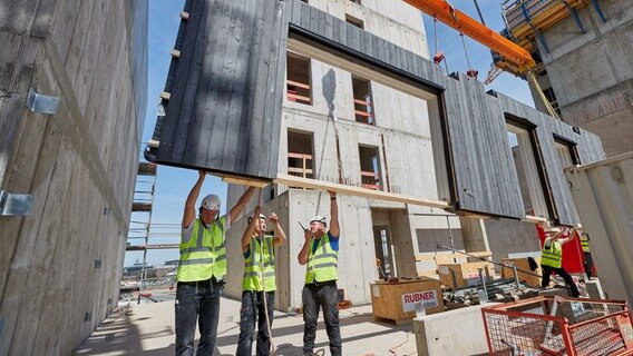 Ein Außenelement aus Holz wird während eines Pressetermins auf der Baustelle mit einem Kran herabgelassen. Das Holzhochhaus «Roots» in der HafenCity bekam seine ersten Holzelemente montiert. Das 65 Meter hohe Gebäude soll 2023 fertiggestellt werden. © picture alliance/dpa | Georg Wendt Foto: Georg Wendt