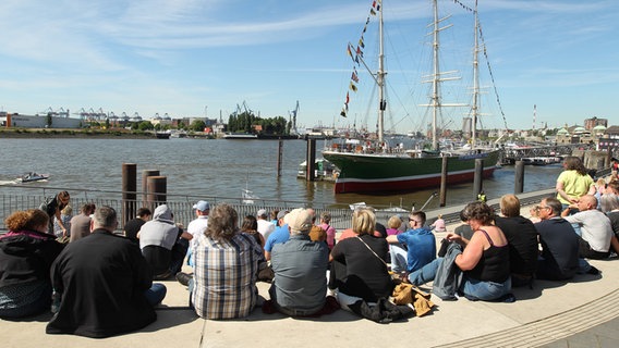 Besucher sitzen an der Jan-Fedder-Promenade. Im Hintergrund das Museumsschiff Rickmer Rickmers. © IMAGO Foto: Hanno Bode