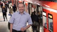 Reinhard Postelt spricht den Hamburg Kommentar. Im Hintergrund sind Fahrgäste beim Ein- und Ausstieg einer S-Bahn zu sehen. © NDR Foto: NDR/Screenshot