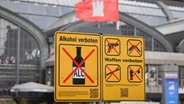 Ein Hinweisschild zum Alkoholverbot ist neben einem Verbotsschild für Waffen am Hachmannplatz vor dem Hauptbahnhof. © dpa Foto: Christian Charisius