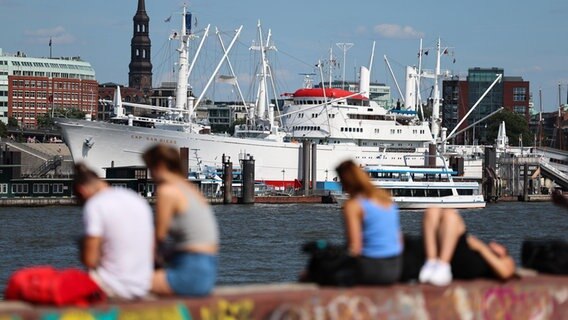 Passanten sitzen bei hochsommerlichen Temperaturen auf einer Mauer am Hafen von Hamburg. © Christian Charisius/dpa 