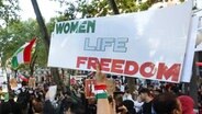 Während einer Demonstration gegen das politische Regime in der Hamburger Innenstadt stehen zahlreiche Teilnehmer mit Fahnen und Schildern zusammen. Auf einem Schild ist die Aufschrift "Women Life Freedom" zu lesen. © picture alliance / dpa Foto: Bodo Marks