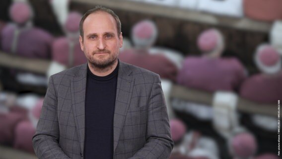 Daniel Kaiser steht vor einem Foto von kirchlichen Würdenträgern. © NDR 