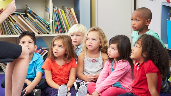 Kleinkinder in einer Büchereri sitzen in einer Gruppe auf dem Fußboden zusammen und gucken zu einer Frau hoch, die etwas vorliest. Von der Frau sieht man nur die Beine. © IMAGO / Shotshop 