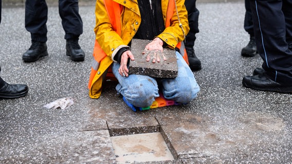 Eine Klima-Aktivistin sitzt mit einem Betonteil an ihrer Hand zwischen Polizisten auf der Fahrbahn der Hamburger Elbbrücken. © picture alliance / dpa Foto: Jonas Walzberg