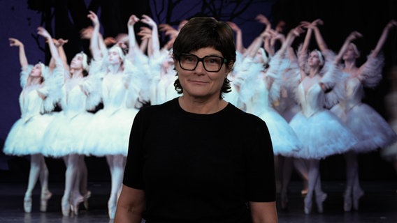 Annette Matz steht vor einem Foto von Balletttänzerinnen auf der Bühne der Hamburger Staatsoper und spricht einen Kommentar zum Abschied des Ballettintendanten John Neumeier. © NDR/Screenshot 