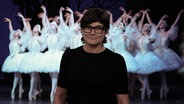 Annette Matz steht vor einem Foto von Balletttänzerinnen auf der Bühne der Hamburger Staatsoper und spricht einen Kommentar zum Abschied des Ballettintendanten John Neumeier. © NDR/Screenshot 