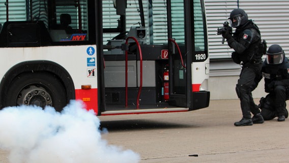 Zwei MKE-Beamte stehen neben einem Bus und haben einen Knaller geworfen, der raucht. © Carolin Fromm/NDR Foto: Carolin Fromm