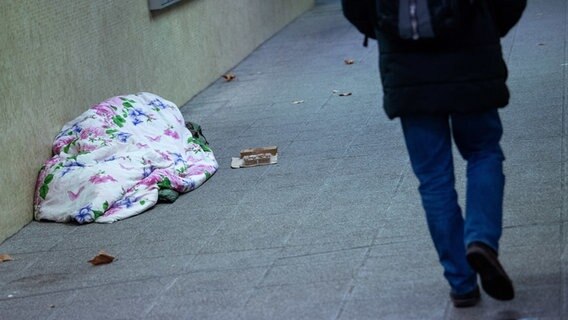 Ein Mensch liegt unter einer Decke auf dem Gehweg, ein anderer Mensch geht vorbei.  Foto: Sebastian Gollnow