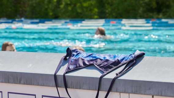 Ein Badeanzug liegt auf der Mauer eines Freibads, dahinter schwimmen Menschen im Becken ihre Bahnen. © picture alliance/dpa | Annette Riedl Foto: Annette Riedl