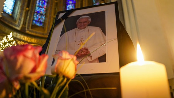 Ein Foto des verstorbenen emeritierten Papsts Benedikt XVI. steht neben Rosen und einer Kerze im Hamburger St. Marien-Dom. Er war im Alter von 95 Jahren im Vatikan gestorben. © picture alliance / dpa Foto: Marcus Brandt