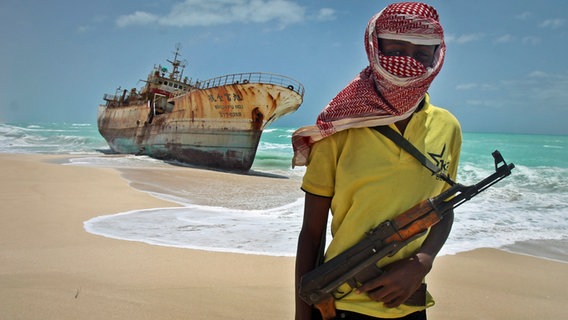 Ein somalischer Pirat steht vermummt und bewaffnet am Strand vor einem verrosteten Tanker. © picture alliance / ASSOCIATED PRESS Foto: Farah Abdi Warsameh