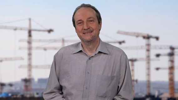 Reinhard Postelt steht vor einem Foto einer großen Baustelle mit mehreren Kränen. © NDR 