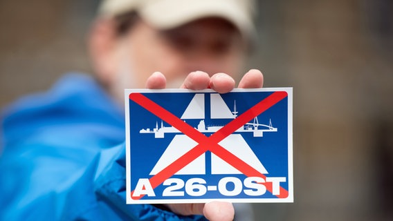 Ein Aktivist des NABU hält während einer Demonstration in Hamburg einen Aufkleber mit dem durchgestrichenen Schriftzug A26-Ost und einem Autobahnsymbol in der Hand. © picture alliance / dpa Foto: Daniel Reinhardt