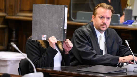 Eine Angeklagte sitzt vor Beginn der Verhandlung im Gerichtssaal neben ihrem Verteidiger. Sie hat ihr Gesicht verdeckt. © picture alliance / dpa Foto: Jonas Walzberg