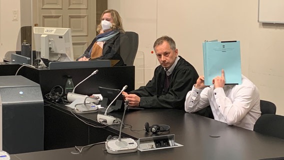 Der Angeklagte sitzt im Gerichtssaal und verdeckt sein Gesicht mit einem blauen Aktenordner. © NDR / Elke Spanner Foto: Elke Spanner