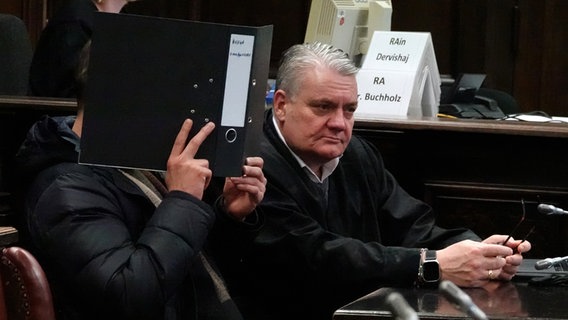 Ein Angeklagter (l.) sitzt neben seinem Anwalt in einem Gerichtssaal in Hamburg und verdeckt sein Gesicht mit einem Aktenordner. Dem 42-Jährigen wird Mord aus niedrigen Beweggründen vorgeworfen. Er soll mit einem Messer auf einen Mann eingestochen haben. © picture alliance / dpa Foto: Rabea Gruber