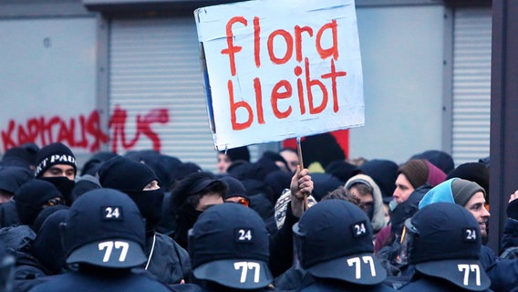 Polizeibeamte stehen am 21.12.2013 vor dem Kulturzentrum «Rote Flora» im Schanzenviertel in Hamburg Demonstranten gegenüber. © dpa Foto: Malte Christians