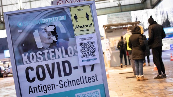 In Hamburg weist ein Schild auf Antigen-Schnelltests hin, daneben warten Menschen. © dpa Foto: Christian Charisius