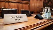 Ein Schild mit dem Text "Olaf Scholz Zeuge" steht auf einem Platz auf der Senatsbank vor Beginn einer Sitzung des Parlamentarischen Untersuchungsausschusses "Cum-Ex" im Plenarsaal der Hamburgischen Bürgerschaft im Rathaus. © picture alliance / dpa Foto: Christian Charisius