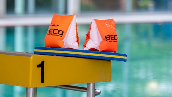 Zwei Schwimmflügel und ein Schwimmbrett liegen auf dem Startblock einer Schwimmhalle. © dpa Foto: David Inderlied