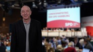 Oliver Wutke steht vor einem Foto, das während eines Parteitags der SPD Hamburg im Bürgerhaus Wilhelmsburg geschossen wurde. © NDR/Screenshot 