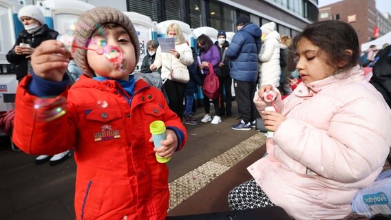Zwei Kinder aus der Ukraine spielen mit Seifenblasen während sie mit ihren Angehörigen vor dem Amt für Migration in Hamburg warten. © dpa Foto: Christian Charisius
