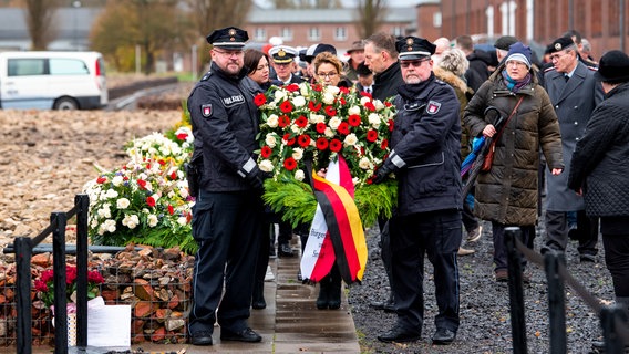 Ein Kranz wird zum Volkstrauertag auf dem Gelände der KZ-Gedenkstätte Neuengamme getragen. Dahinter gehen Hamburgs Zweite Bürgermeisterin Katharina Fegebank (Grünen) und Bürgerschaftspräsidentin Carola Veit (SPD). © dpa Foto: Daniel Bockwoldt