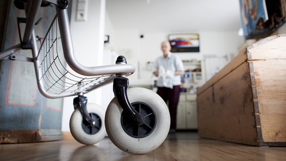 Ein Rollator steht in einer Wohnung, im Hintergrund ist ein älterer Mann zu erkennen. © IMAGO 